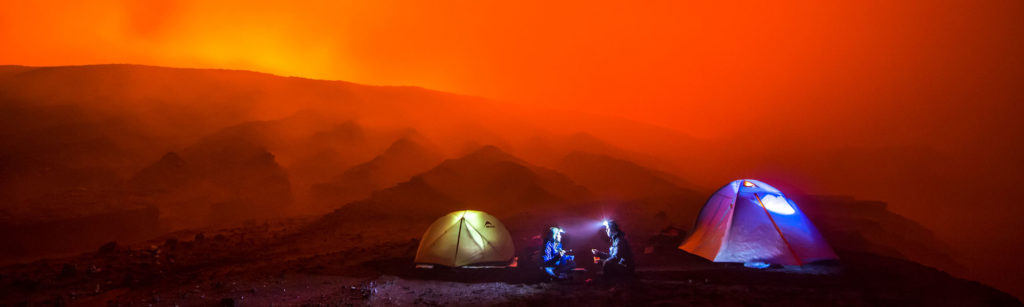 Es ist Nacht, beleuchtete Zelte vor einer rot leuchtendenden Bergkette, der Himmel ist komplett rot.